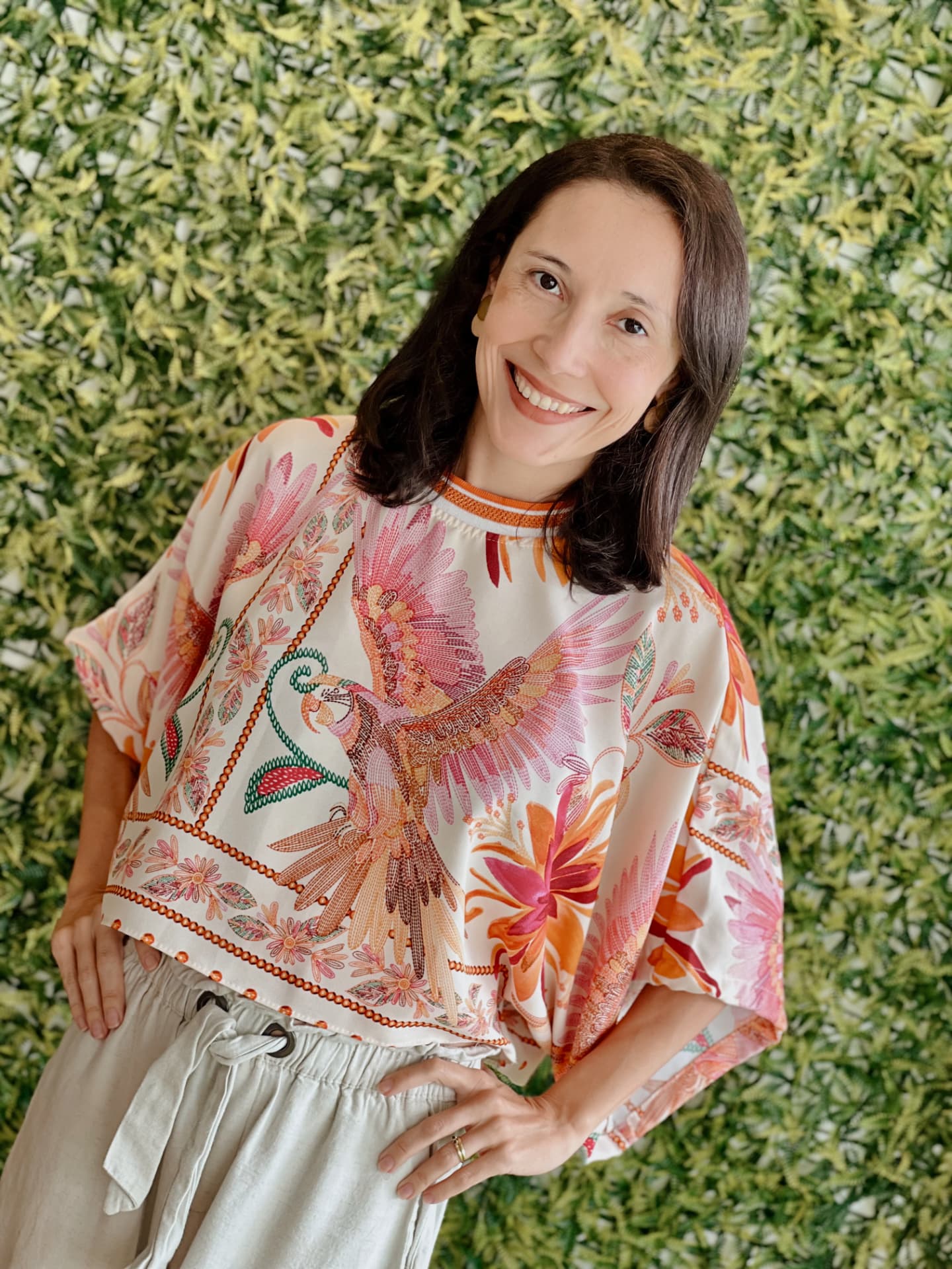 Mari, fundadora da Oca, sorrindo em frente a um jardim vertical, usando blusa floral colorida